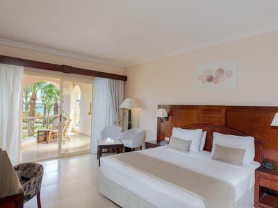 Helles Hotelzimmer mit Doppelbett, Holzkopfteil und Blick auf Terrasse mit Garten.