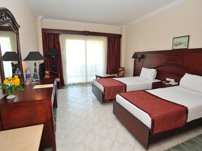 Hotel room with two single beds, wooden furniture, large window, and light tiled floor.
