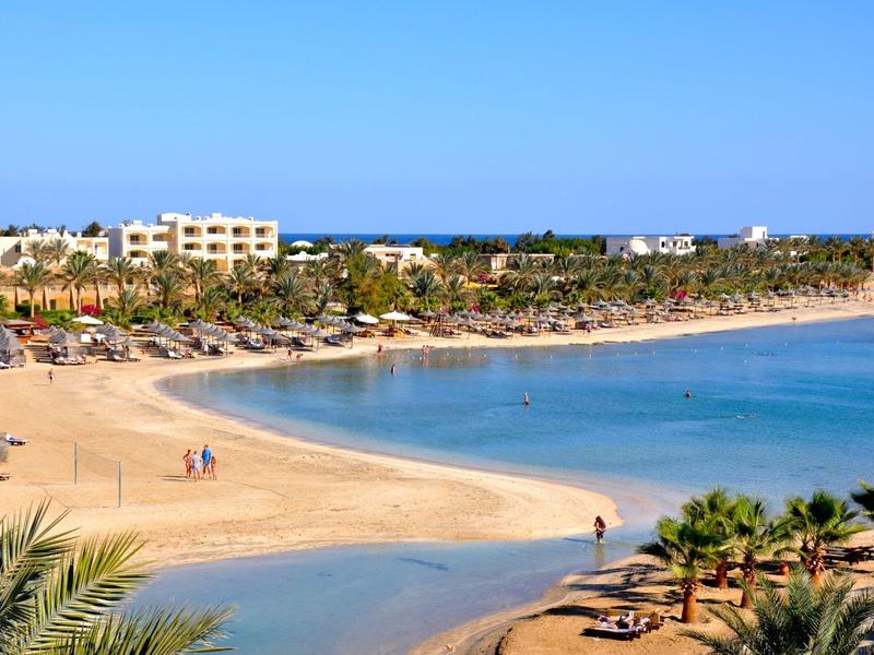Sandy beach with clear water, umbrellas, palm trees, and resort buildings under a blue sky.
