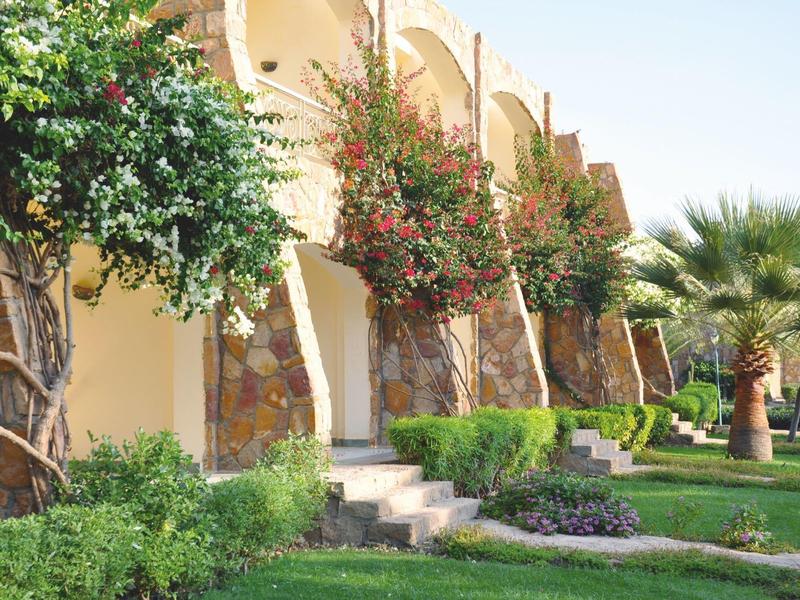 Sunny resort building with stone arches, blooming trees, and green lawn.