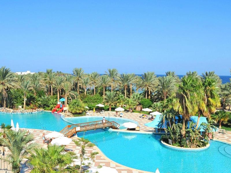 Large outdoor pool with slides, surrounded by palm trees and lounge chairs under clear blue sky.