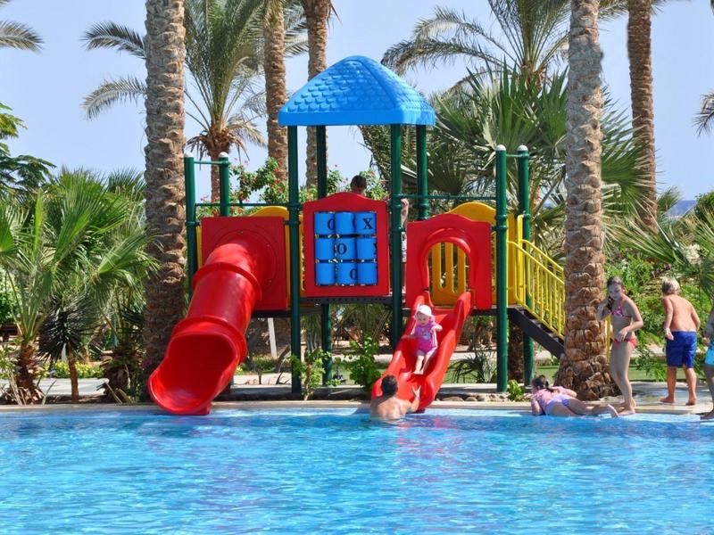Children's water playground with slides next to a swimming pool surrounded by palm trees.