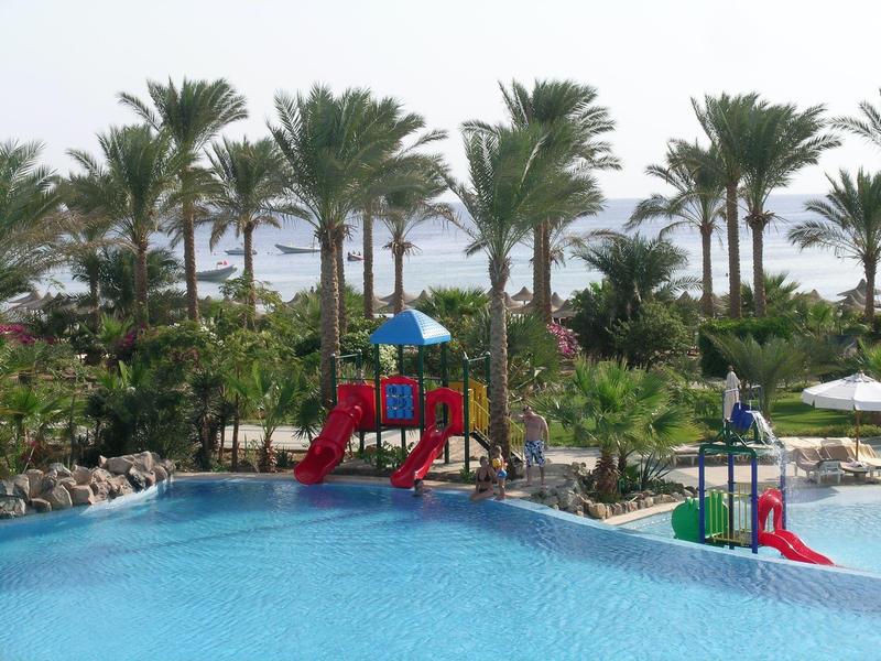 Outdoor pool with kid's slides surrounded by palm trees and tropical plants near the sea.