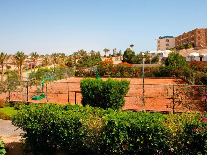 Outdoor tennis court surrounded by greenery and a hotel building under a clear blue sky.