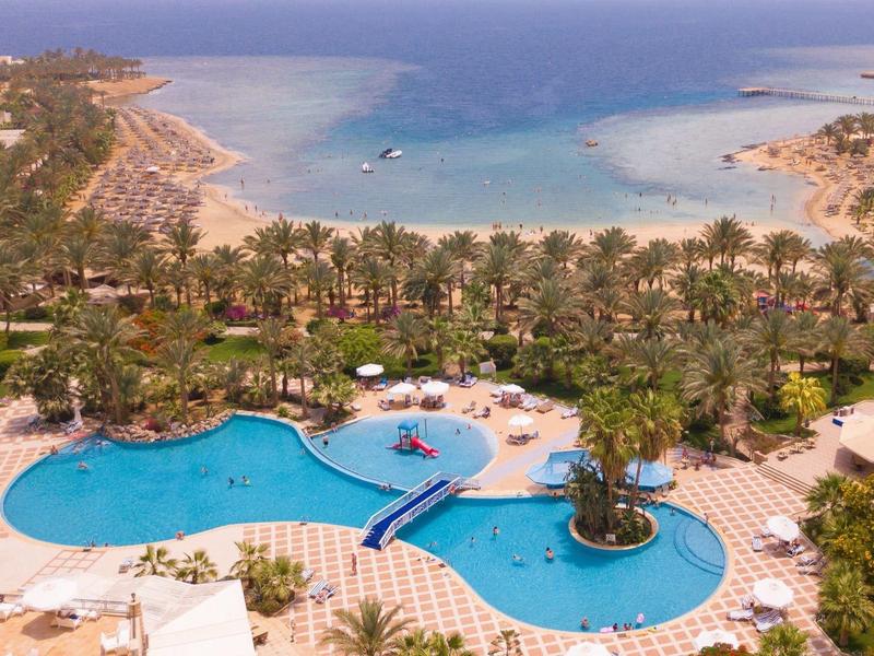 Resort with large curved pool, palm trees, lounge chairs, and beach leading to clear blue sea.