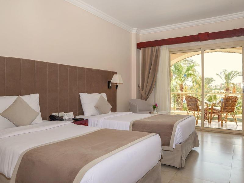 Hotel room with two single beds, beige and white linens, and a balcony overlooking palm trees.