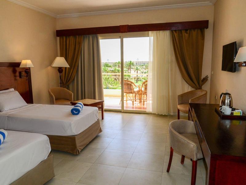 Hotel room with two beds, a balcony with garden view, and a desk with a kettle.
