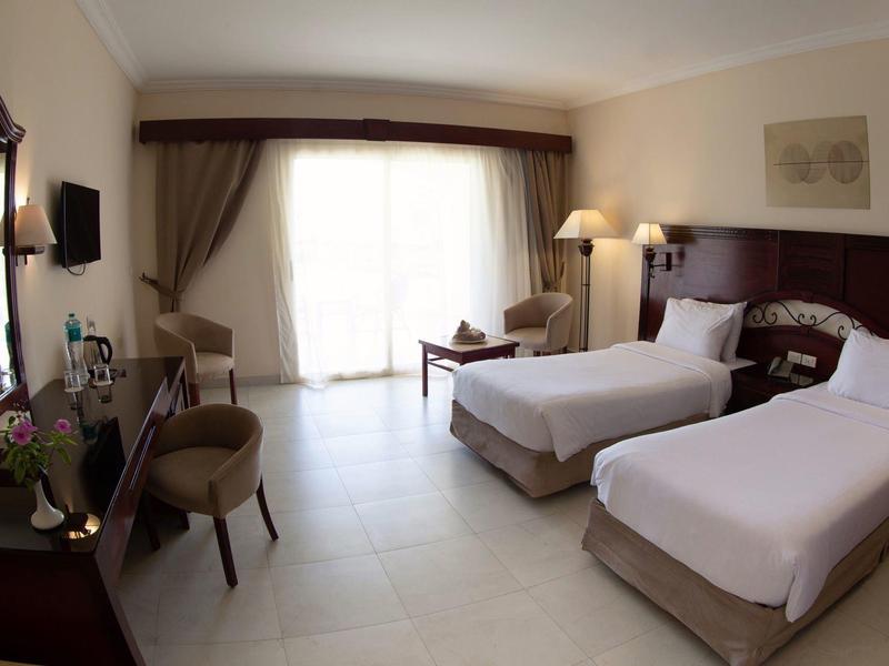 Hotel room with two twin beds, large window with curtains, desk, and chairs in neutral tones.