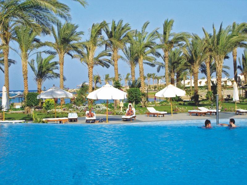 Large outdoor pool with people swimming and sunbathing under palm trees and umbrellas.