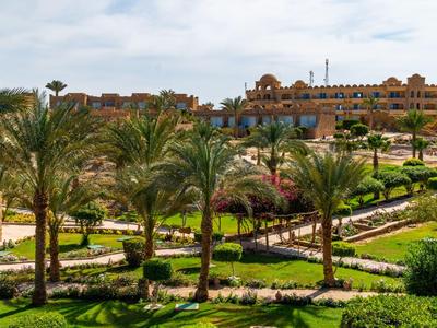 Grüner Hotelgarten mit Palmen und Wegen, im Hintergrund ein großes Gebäude unter blauem Himmel.