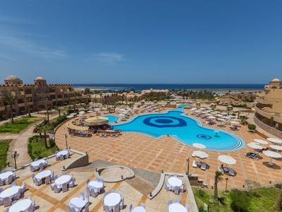 Großer Hotelpool mit Sonnenliegen und Blick auf das Meer unter klarem Himmel.