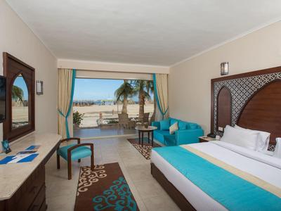 Modern hotel room with double bed, seating area, and view of pool and beach.