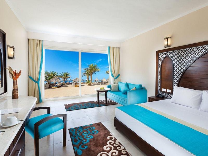 Bright hotel room with large bed, turquoise accents, and sea view through windows.