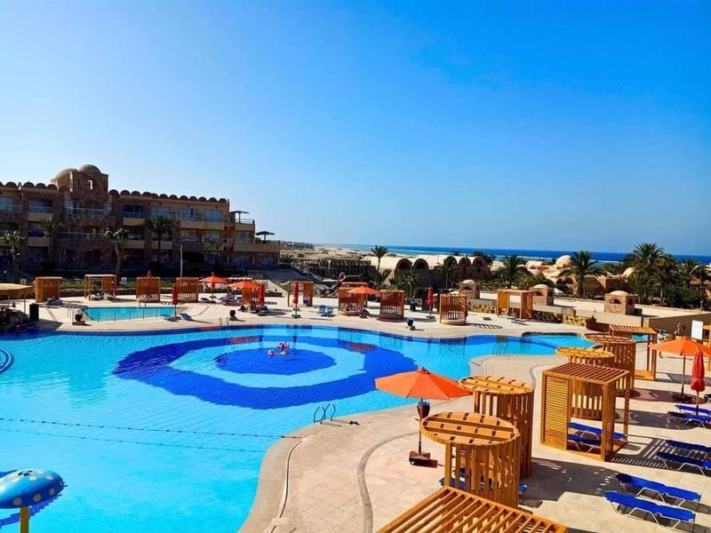 Large hotel pool with blue water patterns, lounge chairs, and orange umbrellas under a clear blue sky.