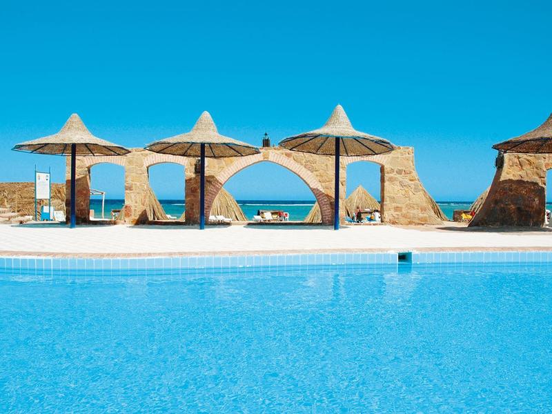 Clear blue pool with straw parasols and seating under a bright blue sky.