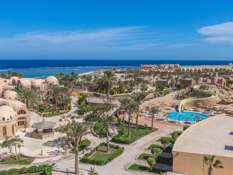 View of a resort with pool, palm trees, and sea in the background under blue sky.