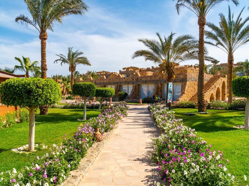 Path through manicured garden with palm trees and flowers leading to hotel building.
