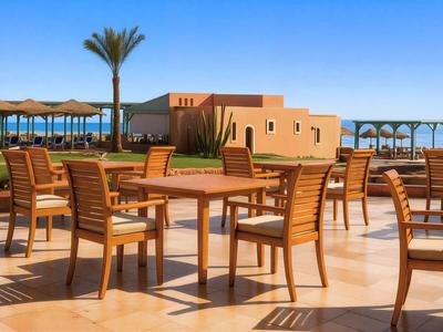 Leerer Außenbereich mit Holztischen und Stühlen, Blick auf Meer und Palmen unter klarem Himmel.