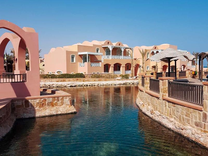 Hotel buildings with pink and beige facades beside a canal under a clear blue sky.