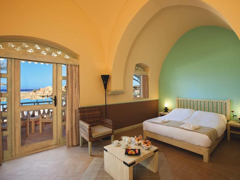 Bright hotel room with double bed, seating area, and balcony overlooking the sea.