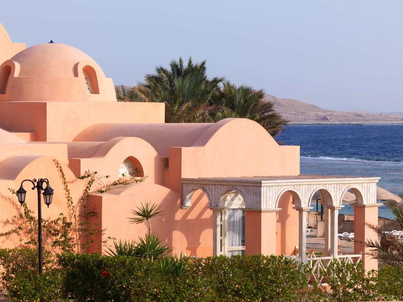 Pink building with domes and arches by the sea with palms in the background.