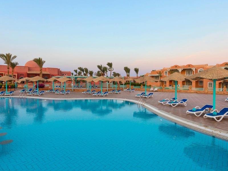 Ein großer Pool mit blauen Liegestühlen und Sonnenschirmen vor einem Hotel bei Sonnenuntergang.