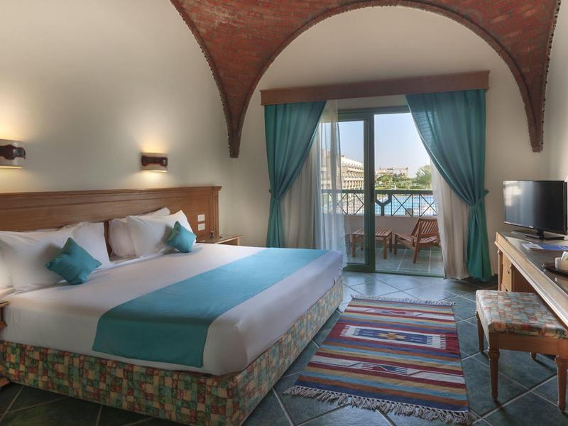 Slaapkamer met tweepersoonsbed, vloerkleden, gordijnen en balkon met zeezicht in een hotel.