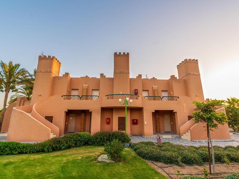 Beige hotelgebouw met torens omgeven door groen en planten bij zonsondergang.
