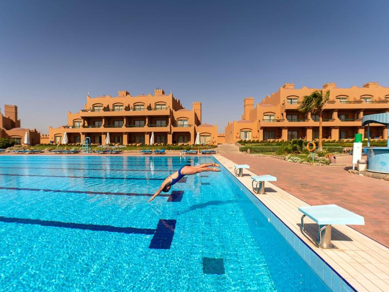 Persoon duikt in een groot, helder buitenzwembad met zonne-terrasen en gebouwen.