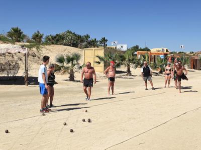 Mehrere Personen spielen Boule auf einem sonnigen Sandplatz mit Palmen im Hintergrund.