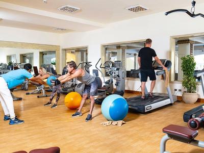 Fitnessraum mit Holzfußboden, Trainingsgeräten, Gymnastikbällen und Menschen beim Workout.
