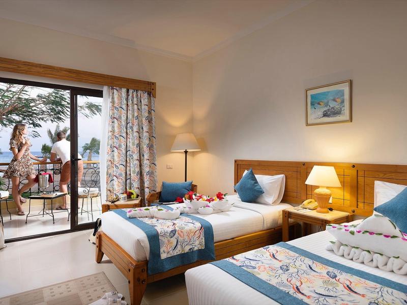 Lichte hotelkamer met twee eenpersoonsbedden, houten meubels en balkon met uitzicht op bomen.