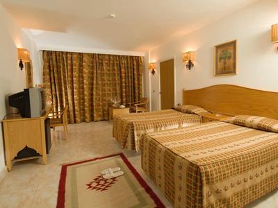 Hotelzimmer mit zwei Einzelbetten, Holzmöbeln, Teppich und goldfarbenen Vorhängen.