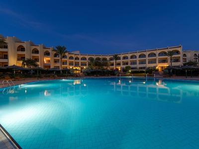 Beleuchtetes Hotel mit großem Pool und Palmen bei Nacht unter dunkelblauem Himmel.