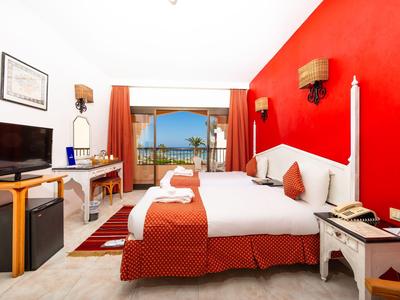 Hotelzimmer mit roter Akzentwand, zwei Betten und Balkon mit Meerblick.