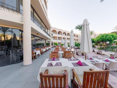 Terrassenrestaurant mit Holztischen, weißen Tischdecken und Stühlen, Sonnenschirm und Pflanzen.