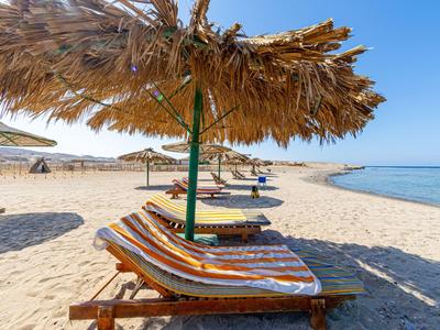 Strand mit Strohschirm und Liegen auf hellem Sand, klarer blauer Himmel und ruhiges Meer im Hintergrund.