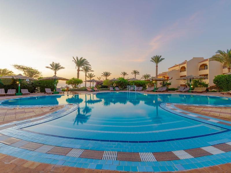 Großer sauberer Pool mit blauem Wasser, umgeben von Palmen und beigefarbenen Gebäuden im Sonnenuntergang.