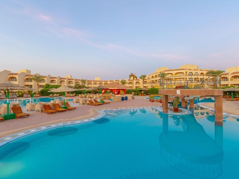 Großer Pool mit blauer Wasseroberfläche, umgeben von Sonnenliegen und gelben Hotelgebäuden bei klarem Himmel.