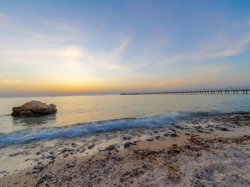 Strand mit Sand, Steinen, ruhigem Meer und blau-orangefarbenem Himmel bei Sonnenuntergang.