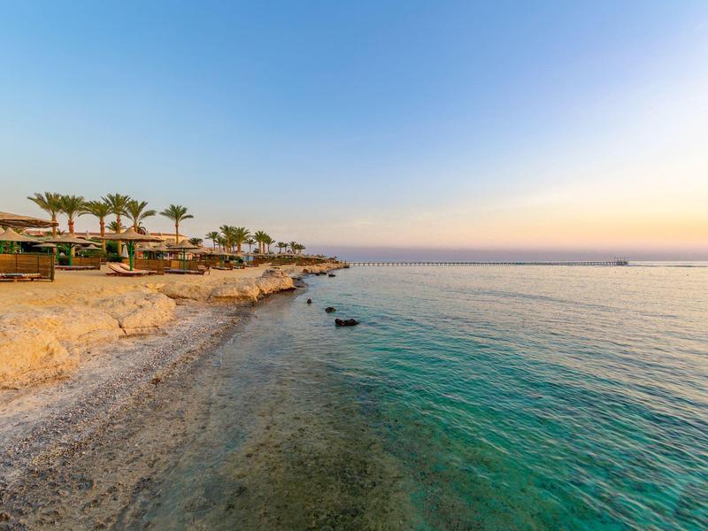Klares Meer mit türkisfarbenem Wasser, Felsenstrand und Palmen unter blauem Himmel bei Sonnenuntergang.