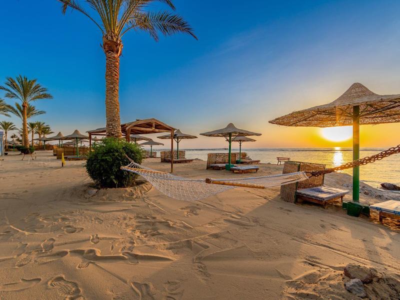 Strand mit Palmenschirm, Sonnenliegen, Palmen und Sonnenuntergang am ruhigen Meer.