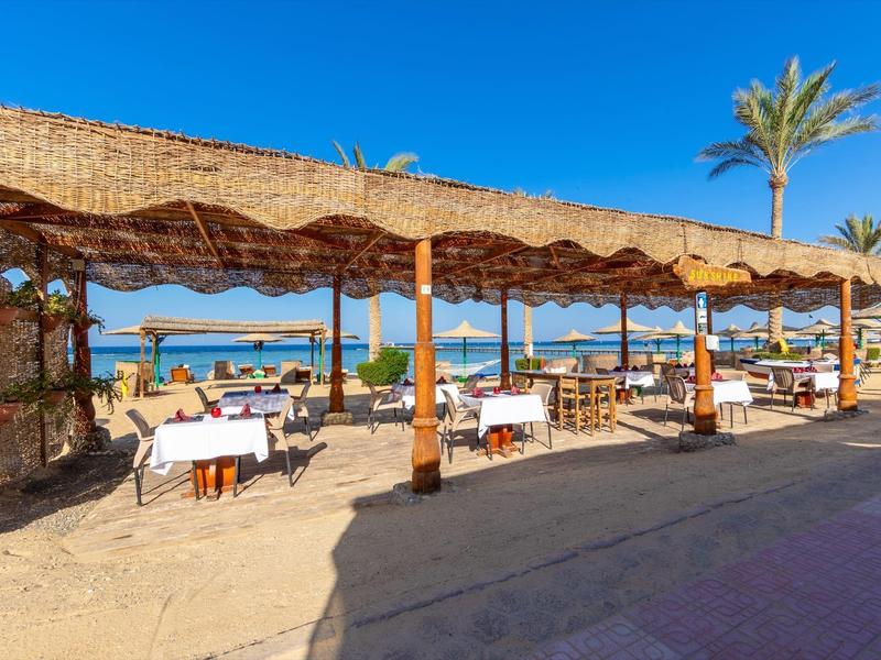 Offenes Strandrestaurant mit Holzmöbeln, weißen Tischtüchern und Blick aufs blaue Meer bei Sonnenschein.