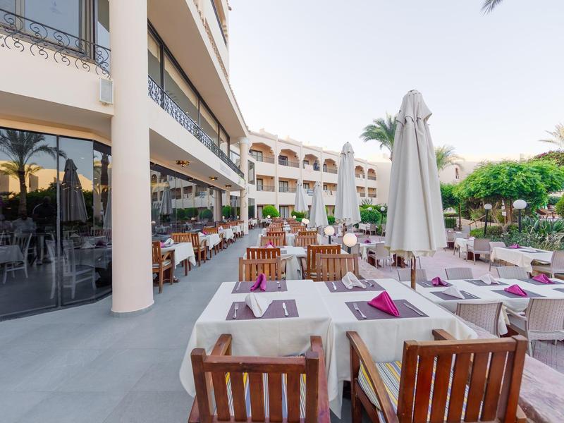 Terrassenrestaurant mit Holztischen, weißen Tischdecken und Stühlen, Sonnenschirm und Pflanzen.