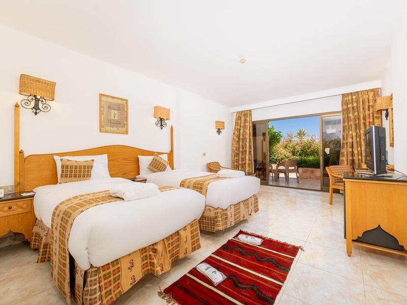 Helles Hotelzimmer mit zwei Einzelbetten, Holzmöbeln und Balkon mit Blick auf Palmen.