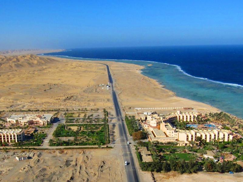 Blick auf Küstenstraße mit Gebäuden und Gärten in Wüstenlandschaft neben blauem Meer.