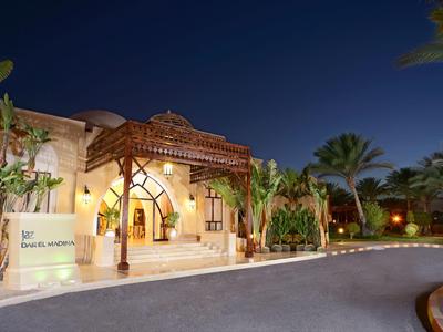 Eingang eines Hotels bei Nacht mit beleuchteter Fassade, Palmen und tropischer Vegetation.