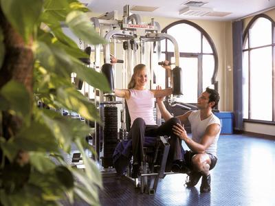 Frau trainiert an Fitnessgerät, Trainer gibt Anweisungen, heller Raum mit großen Fenstern.