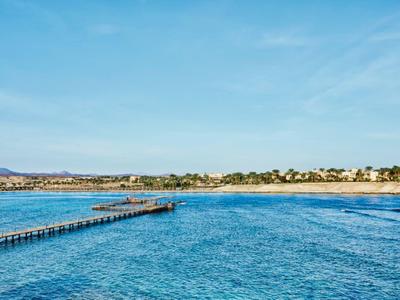 Breiter Steg ragt in ruhiges blaues Meer, Strand mit Häusern und klarem Himmel im Hintergrund.
