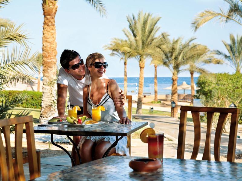 Couple enjoying drinks at a resort patio with palm trees and a sea view.